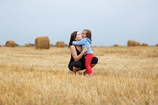 Mother and daughter hugging in the middle sloping field on straw bales background. Shallow depth of field. Selective focus on the models.