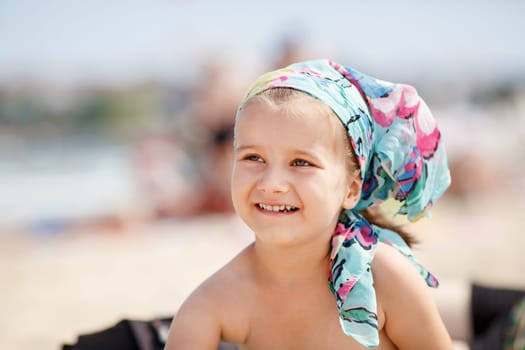 A smiling little girl child with a colorful handkerchief on his head. Hot sunny summer day at the beach. Selective focus on child's face.