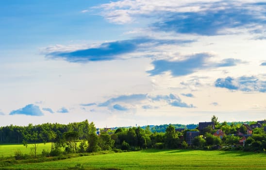 Sunny summer evening. Rural landscape. The sky with clouds, grass field, trees and farmhouses.