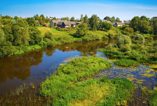 River with thickets of trees and shrubs along the banks. Sunny summer landscape. Calm surface of the water with the reflection of blue sky and vegetation.