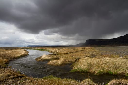 Horizontal panorama view of an Icelandic river running through the grass with rain stormy clouds above and mountains on the right side