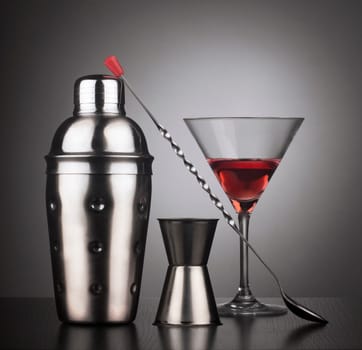 Cocktail tools with shaker, spoon and a glass filled with red liquid,  square composition