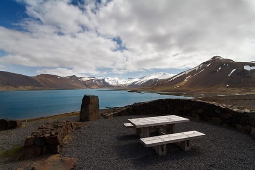 Horizontal panorama of a wooden resting bench next to a lake and mountains covered in snow in Iceland