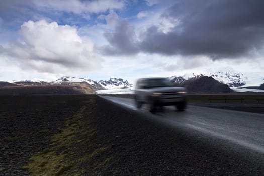 Horizontal photo of an off road car on a straight asphalt road coming from the mountains with clouds above and the Vatnajokull glacier in the background, Iceland
