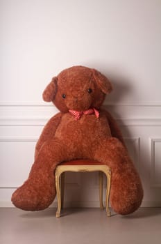 Huge toy bear with a dotted bow standing on a chair against a white wall