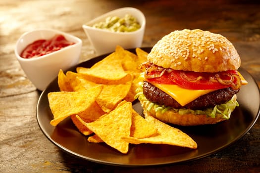 Delicious large cheeseburger topped with tomatoes and onions alongside triangular nacho chips and bowls of salsa and guacamole