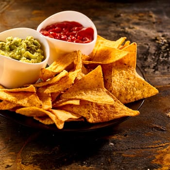 Plate full of guacamole, salsa and nacho chips close up on old worn out dark stained table