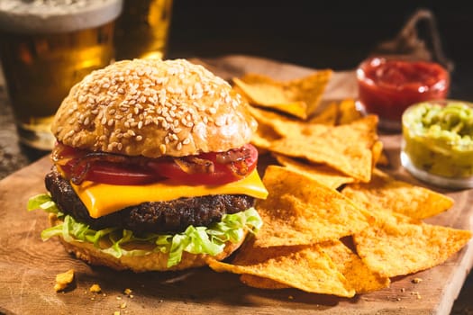 Large freshly prepared cheeseburger topped with tomatoes, onions and lettuce with nacho chips, salsa, guacamole and beer in background