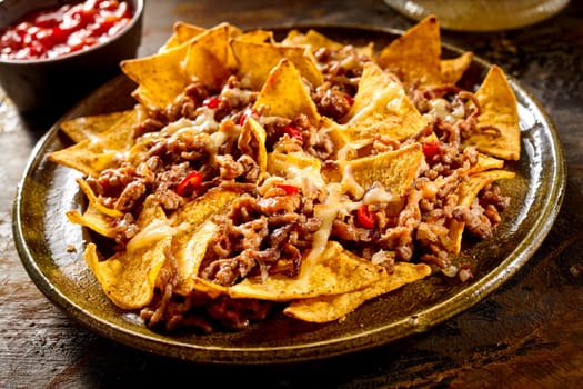 Plate full of yellow corn nachos and cooked ground beef with bowl of red salsa in background over wooden table