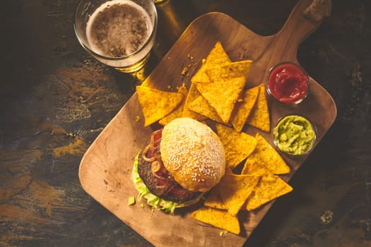 Top down view on burger in sesame bun, tortilla chips, salsa and guacamole on cutting board with beer on table