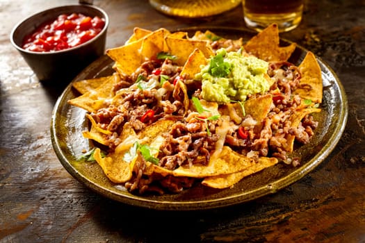 Yellow corn nacho chips garnished with ground beef, guacamole, melted cheese, peppers and cilantro leaves in plate on wooden table