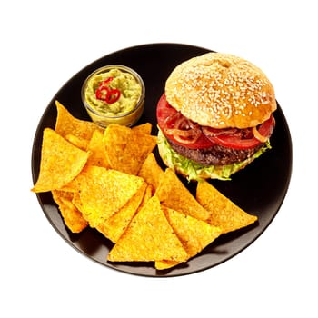 Top down view on isolated round black plate of triangular tortilla chips, guacamole and burger in sesame seed bun over white background
