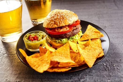Delicious cheeseburger topped with tomato, fried onions and lettuce in sesame bun, tortilla chips, guacamole beside two glasses of beer
