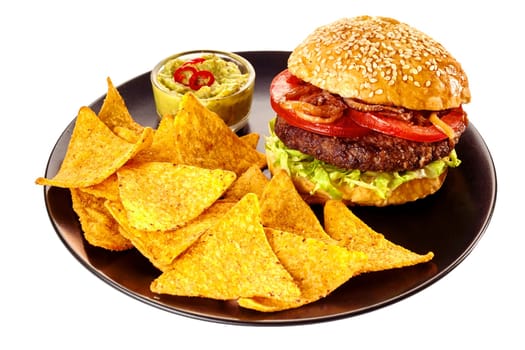 Isolated round black plate of nacho tortilla chips, guacamole and hamburger topped with tomatoes and friend onions in sesame seed bun over white background