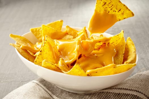 Single triangular yellow corn tortilla chip pulled out of bowl of cheese covered nachos over gray and white tablecloth