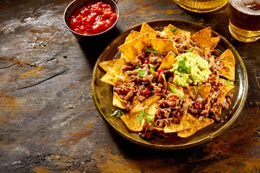 Delicious plate of yellow corn tortilla chips with cheese, meat, guacamole and red hot spicy salsa over table with copy space