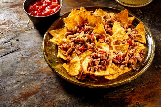 Plate full of yellow corn tortilla chips, melted cheese and cooked ground beef with bowl of red salsa in background over wooden table with copy space