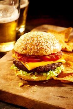Single delicious well done burger topped with onions, tomatoes, cheddar cheese slice and lettuce with nacho chips on cutting board and two glasses of beer in background