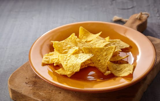 Single plate of tasty yellow tortilla chips on thick wooden cutting board over gray table with copy space