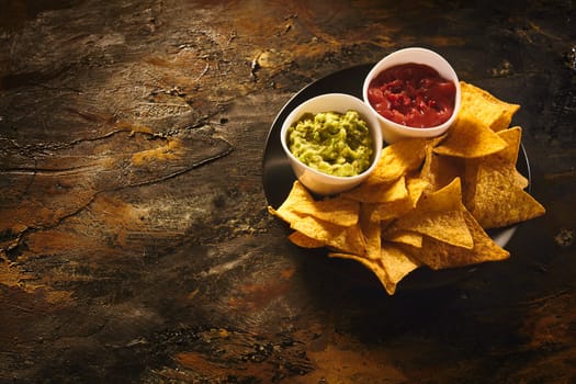 Top down view on delicious crunchy appetizer of triangular yellow corn tortilla chips beside little bowls of guacamole and salsa dip over old dark wooden table with copy space