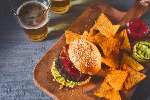 Cropped view on freshly cooked burger in sesame bun, tortilla chips, salsa and guacamole on cutting board with beer on table