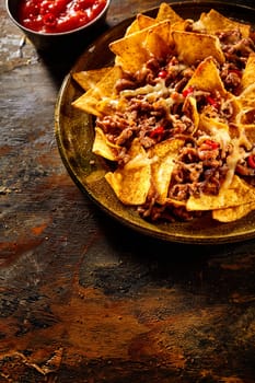 Plate full of yellow corn nachos, melted cheese and cooked ground beef with bowl of red salsa in background over wooden table with copy space