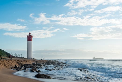 DURBAN, SOUTH AFRICA - MARCH 12, 2016: Umhlanga Rocks Lighthouse with fishermen and people walking on the promenade. Ship on the Indian Ocean in the background