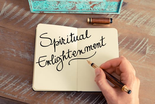 Handwritten text Spiritual Enlightenment as concept image. Retro effect and toned image of a woman hand writing a note with fountain pen on a notebook.