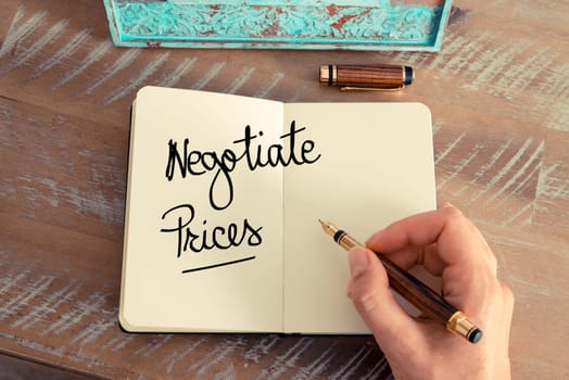Handwritten text Negotiate Prices as concept image. Retro effect and toned image of a woman hand writing a note with fountain pen on a notebook.