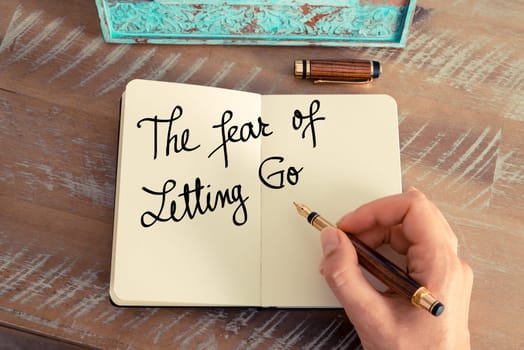 Handwritten text The Fear Of Letting Go as concept image. Retro effect and toned image of a woman hand writing a note with fountain pen on a notebook.