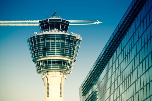 Flights management air control tower and passenger terminal in Munich international airport with flying plane in clear sky. Stock photo with split toning effect.