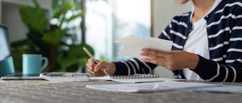 A person calculates their income and bills using documents, a calculator, and a notebook on a table at home.