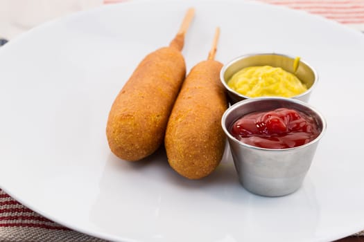 Two corn dogs with metal condiment cups containing mustard and ketchup on a white plate.