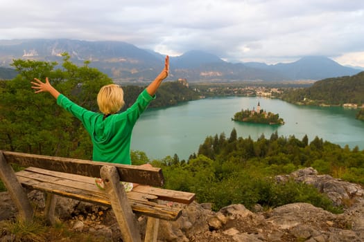 Relaxed female traveler, arms rised, enjoying beautiful panoramic view of Julian Alps, Lake Bled with St. Marys Church of the Assumption on the small island, Bled, Slovenia, Europe.