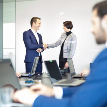 Sealing a deal. Business people shaking hands, finishing up meeting in corporate office. Businessman working on laptop in foreground. Business and entrepreneurship concept.