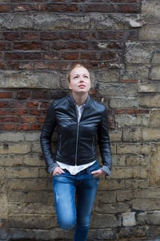 Fashion street style portrait of young beautiful calm female model, hands in pockets, leaning on vintage brick wall in background.