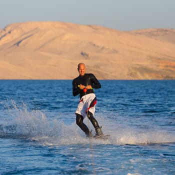 Wakeboarder in wetsuit riding in sunset. Wakeboarding is a surface water sport which involves riding a wakeboard over the surface of a body of water.