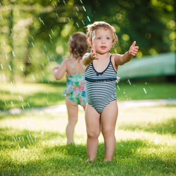 The two little baby girls playing with garden sprinkler. Summer outdoor water fun and green grass.
