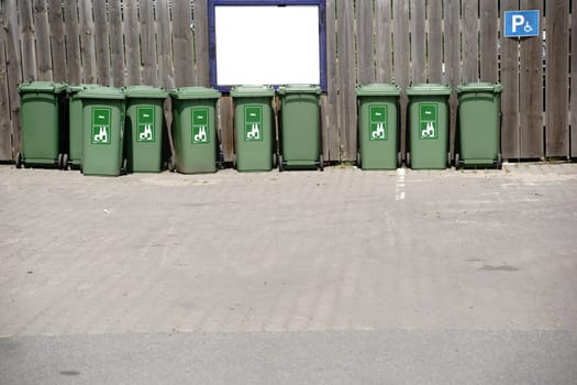 A row of green dumpsters for glass waste stands in front of a board fence.