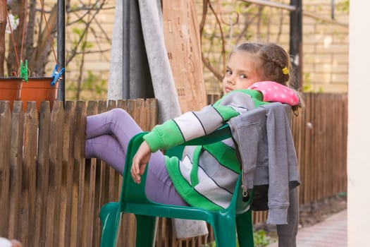 Seven-year girl is resting sitting on a chair in the yard and putting his feet on the wooden fence