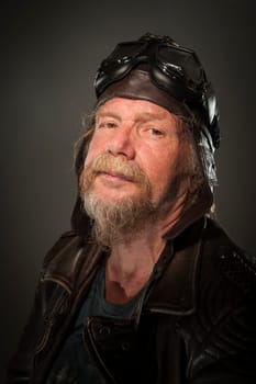 lateral head-and-shoulder portrait of an older man with a full bee,leather jacket and motorcycle goggles looking seriously into the camera