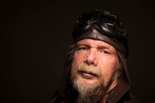lateral head-and-shoulder portrait of an older man with a full bee,leather jacket and motorcycle goggles looking seriously into the camera