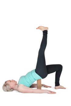Full body view of a young blonde woman during yoga exercise "half bridge with her leg stretched upwards"" against a white background with a slight shadow. Wooden block as assistance under the buttocks."