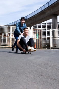 Young couple having fun in the city. The woman is pushing her partner along as he sits on a skateboard. They are both laughing.