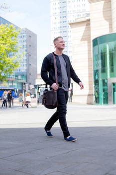 Young man walking in the city. He has a leather bag and is dressed in casual clothing.