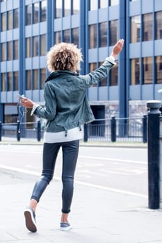 Young woman signalling for a taxi in the city centre. She is holding a smartphone with headphones and has her hand in the air.