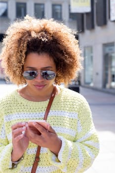 Woman in the city. She is wearing sunglasses and looking down to her smartphone which she is holding.