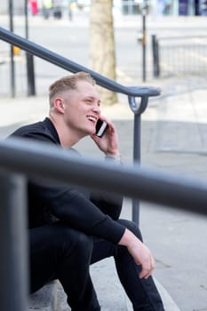 Young man sitting on some steps in the city. He is using his smartphone to have a phonecall.