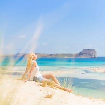 Relaxed woman enjoying sun, freedom and life an a beautiful sandy beach of Balos in Greece. Young lady feeling free, relaxed and happy. Vacations, freedom, happiness, enjoyment and well being.