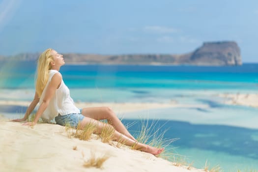Relaxed woman enjoying sun, freedom and life an a beautiful sandy beach of Balos in Greece. Young lady feeling free, relaxed and happy. Vacations, freedom, happiness, enjoyment and well being.